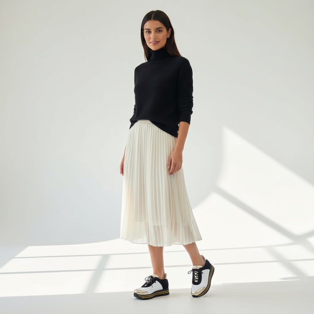 Woman wearing a black turtleneck, white pleated skirt, and black and white sneakers in a bright room.
