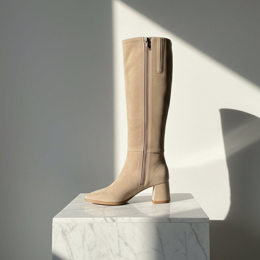 A tall knee-high beige suede boot with a side zipper, block heel, and pointed toe, photographed standing upright on a white surface against a white wall.