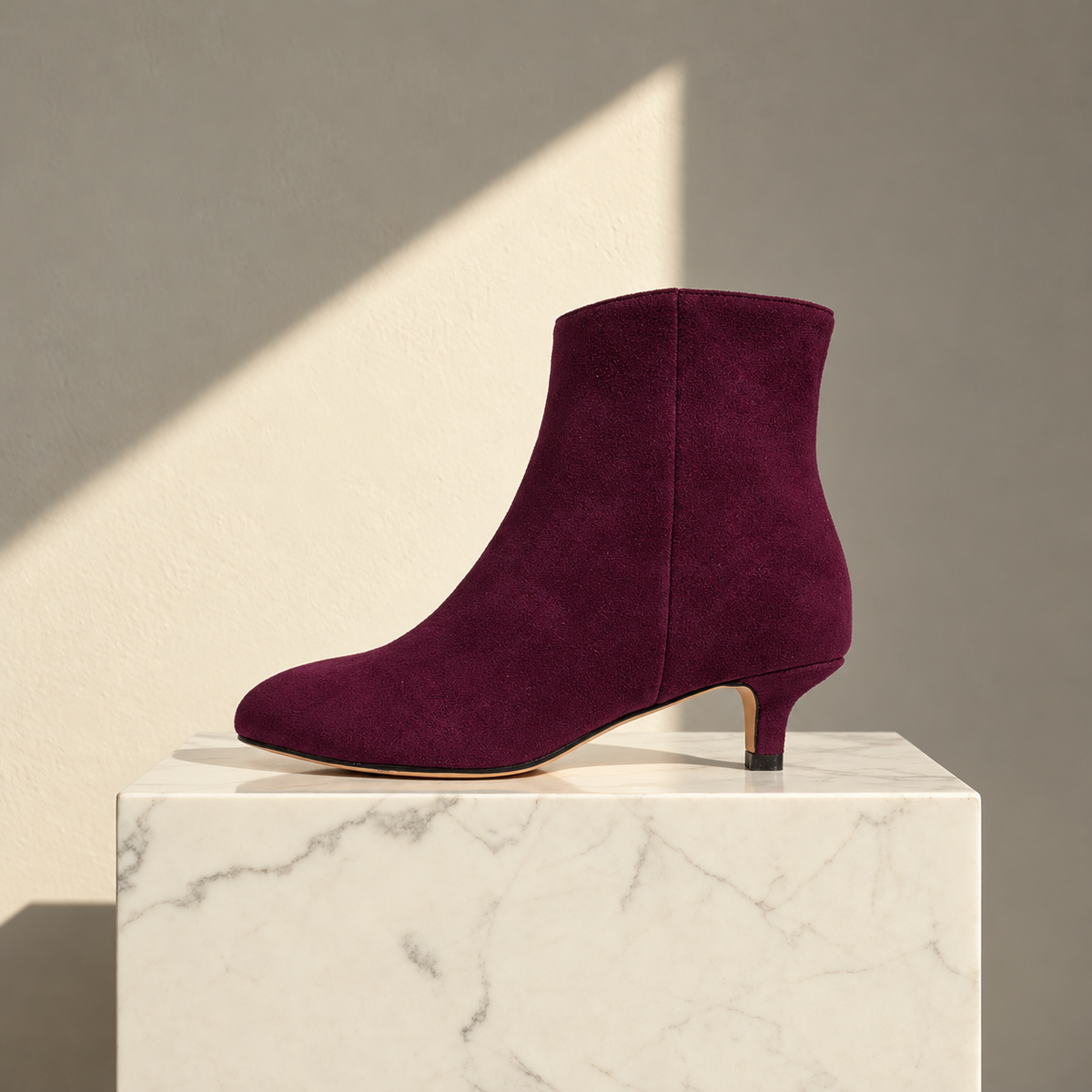 A single ankle boot in deep burgundy suede with a kitten heel, displayed on a white marble pedestal against a warm gray wall with a diagonal shadow.