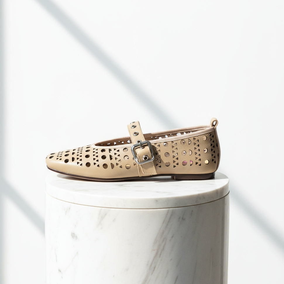 Beige Mary Jane flats with geometric cutouts displayed on marble with tropical leaves.
