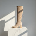 A single knee-high boot in light beige suede or nubuck, standing on a white marble pedestal against a gray wall with sharp window-light shadows.