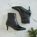 Black high-heeled ankle boots on a marble surface with a green plant.