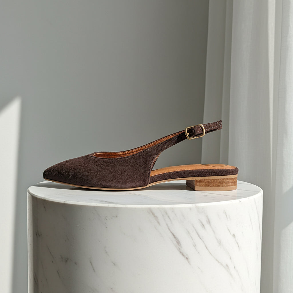 Brown suede pointed toe ballerina slingbacks with a gold buckle set on a cylinder shape marble stand.