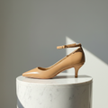 A single tan leather pointed-toe pump with a kitten heel and ankle strap, displayed on a white marble cylindrical pedestal against a neutral gray background with sharp sunlight shadows.