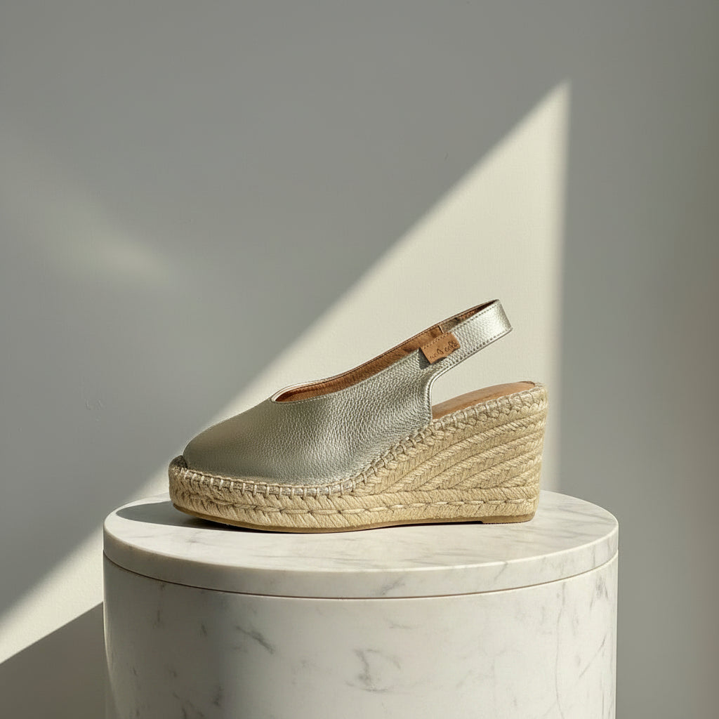 A metallic silver espadrille wedge shoe with a slingback strap, displayed on a white marble cylinder pedestal against a gray wall with a diagonal shadow.