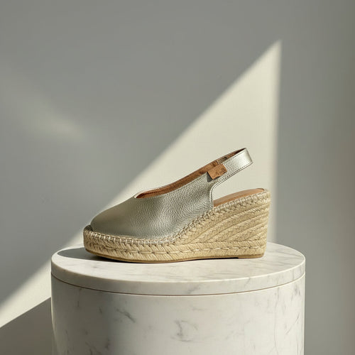 A metallic silver espadrille wedge shoe with a slingback strap, displayed on a white marble cylinder pedestal against a gray wall with a diagonal shadow.