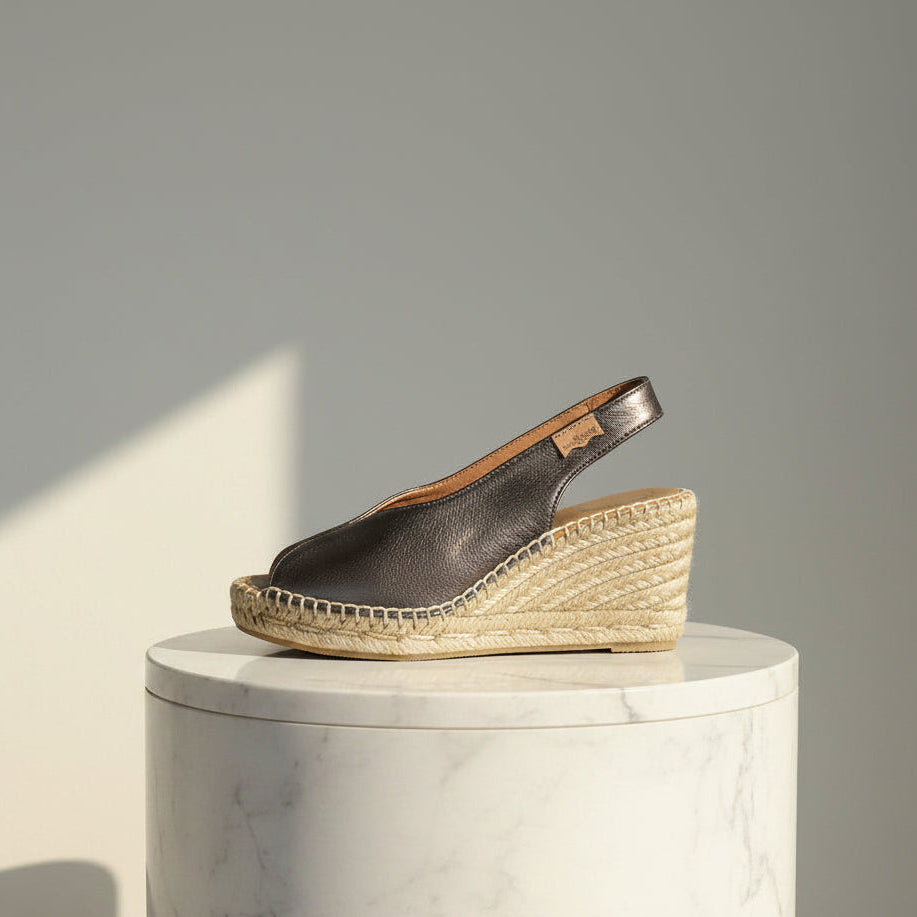 A single wedge espadrille sandal in dark brown leather with a natural jute-wrapped wedge heel, displayed on a white marble cylinder pedestal against a neutral gray background.