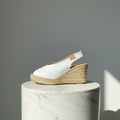 A single women's wedge espadrille sandal with a white leather upper and a natural jute-wrapped wedge heel, photographed from the side against a textured gray background.