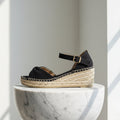 Black wedge sandal with jute sole on a white marble pedestal against a light gray background