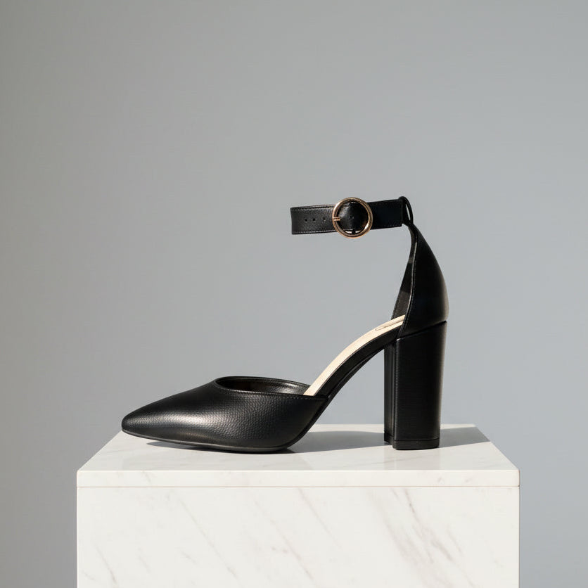 Black high-heeled shoe with ankle strap on a white pedestal against a gray background