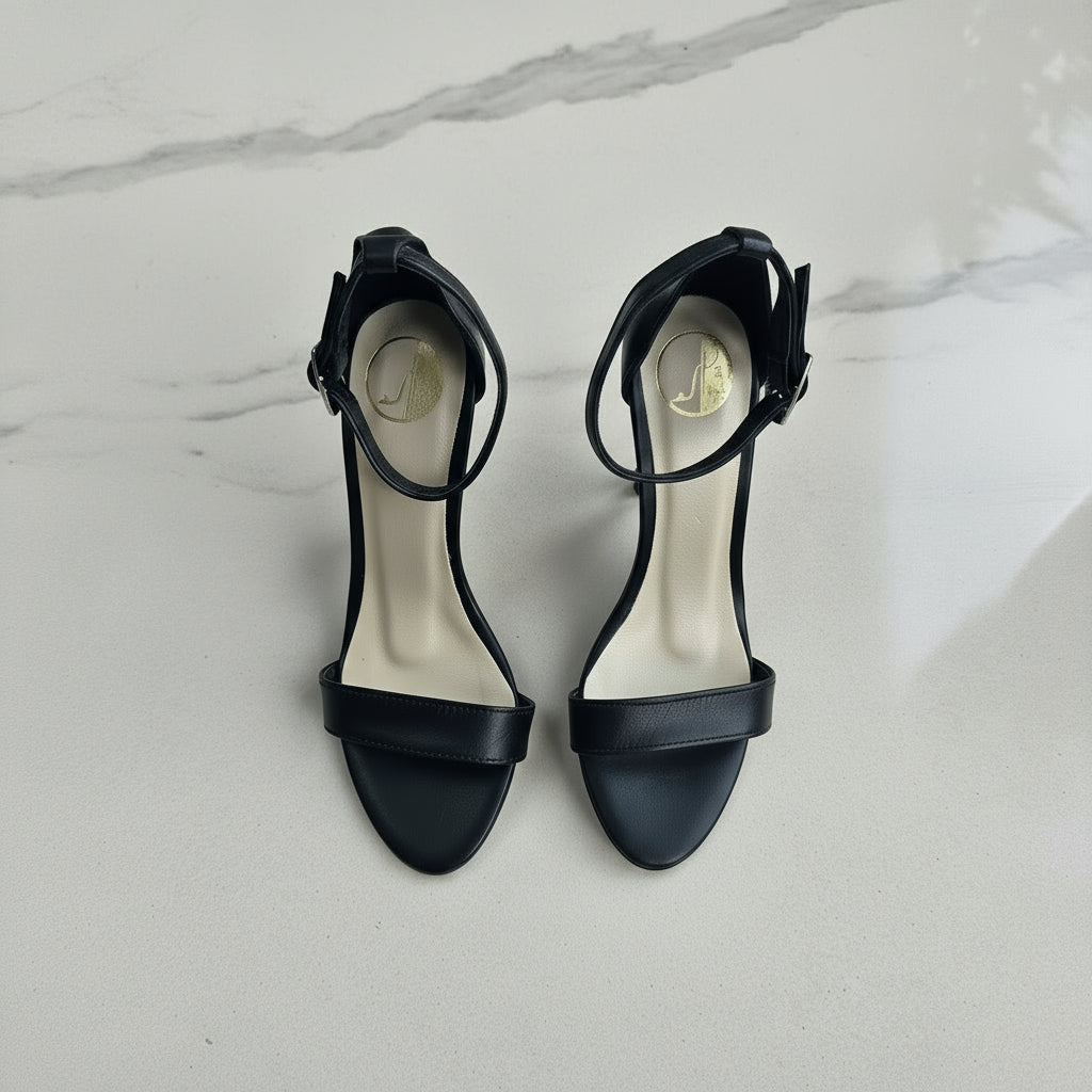 Black high-heeled sandals on a marble surface with green leaves.