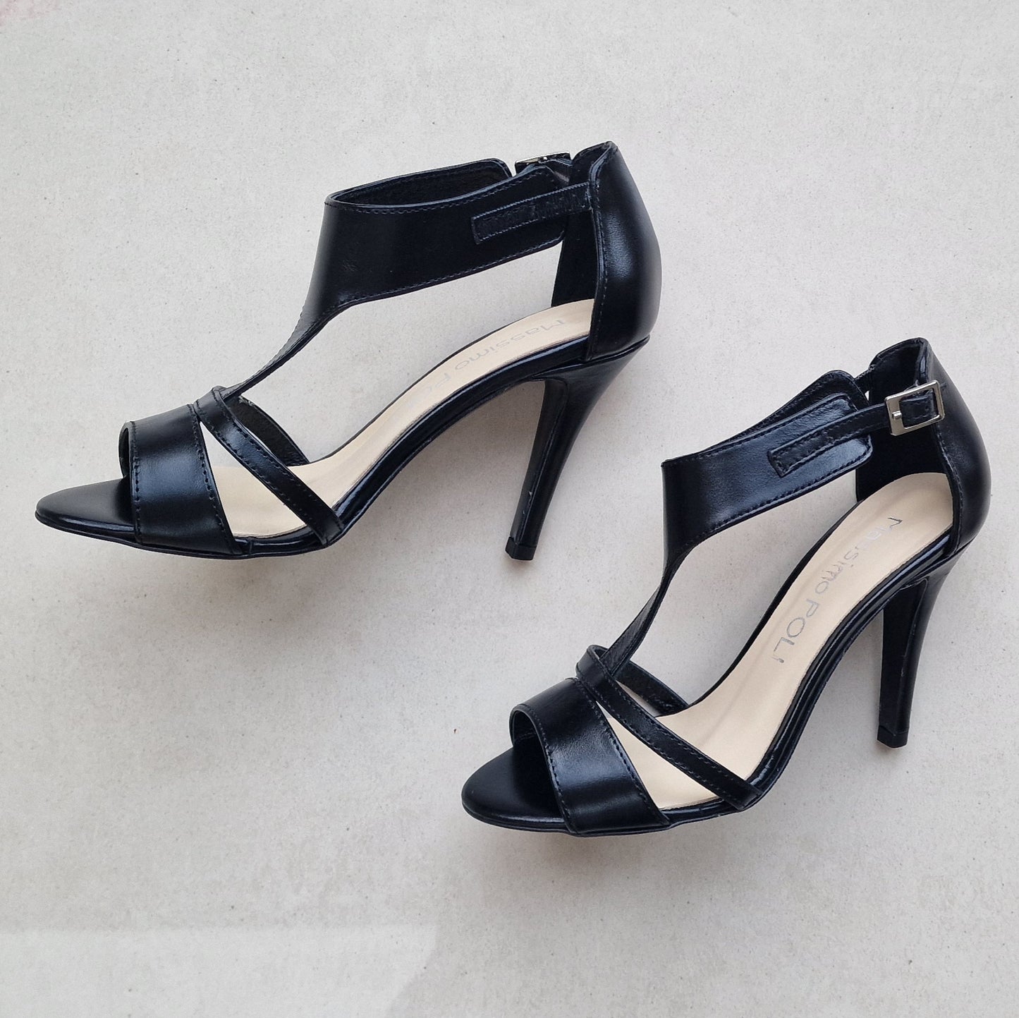 Black leather high heel sandals with a silver buckle lying on a marble floor. 