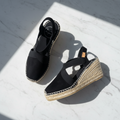 A pair of black wedge espadrille sandals with elastic straps and braided jute-wrapped wedge heels, photographed on a white marble surface with strong diagonal sunlight casting shadows.