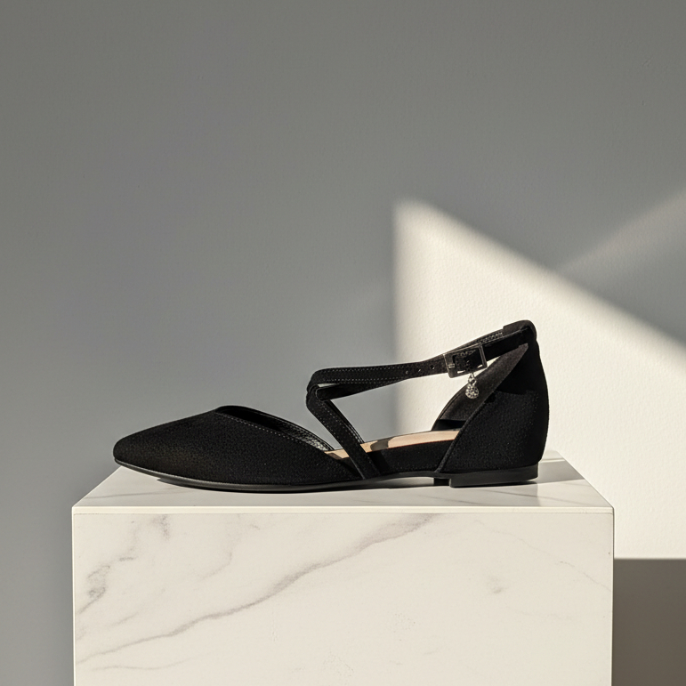 A single black flat sandal with a pointed toe, T-strap design, and ankle buckle, displayed on a white marble pedestal against a gray background with a strong diagonal shadow.