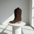 Brown cowboy boot on a white pedestal against a light gray wall with sunlight casting shadows.