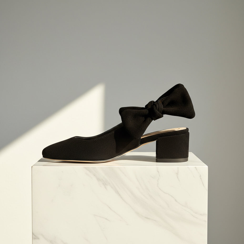 A pair of black suede slingback pumps with a block heel and a bow detail on the strap, displayed on a white cardboard box.