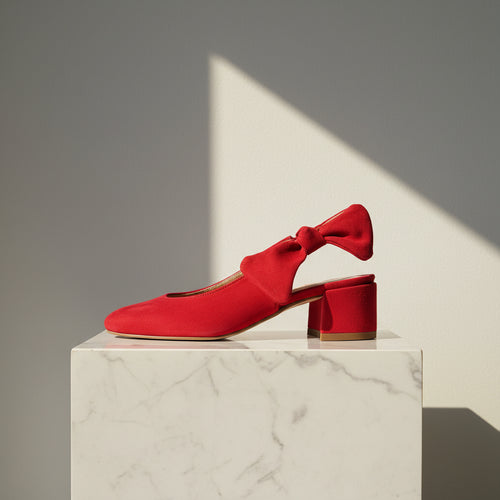 A bright red slingback mule shoe with a bow detail and block heel, displayed on a white marble pedestal against a gray background with dramatic diagonal lighting.