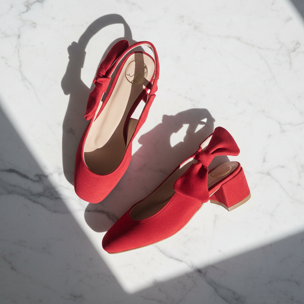 A pair of red pointed-toe slingback pumps with bow details and block heels, styled on a white marble surface with dramatic diagonal shadows.