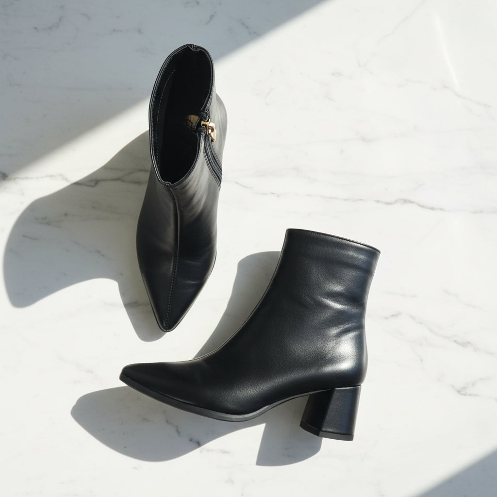 Pair of CORA petite black leather pointed-toe ankle boots with gold zip pull and block heel, laid flat on white marble in natural sunlight.