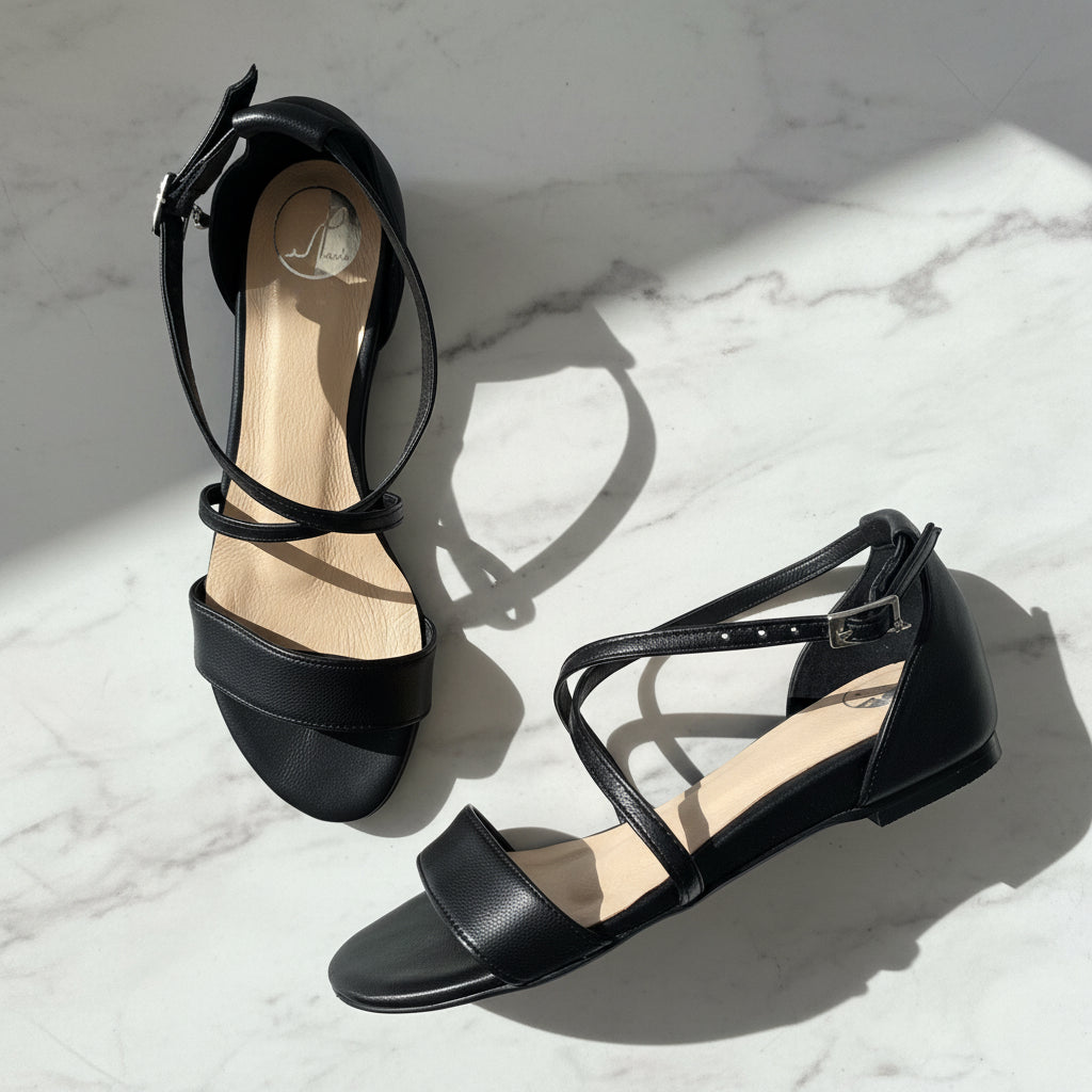 Pair of black flat sandals with cross straps set on a marble surface. 