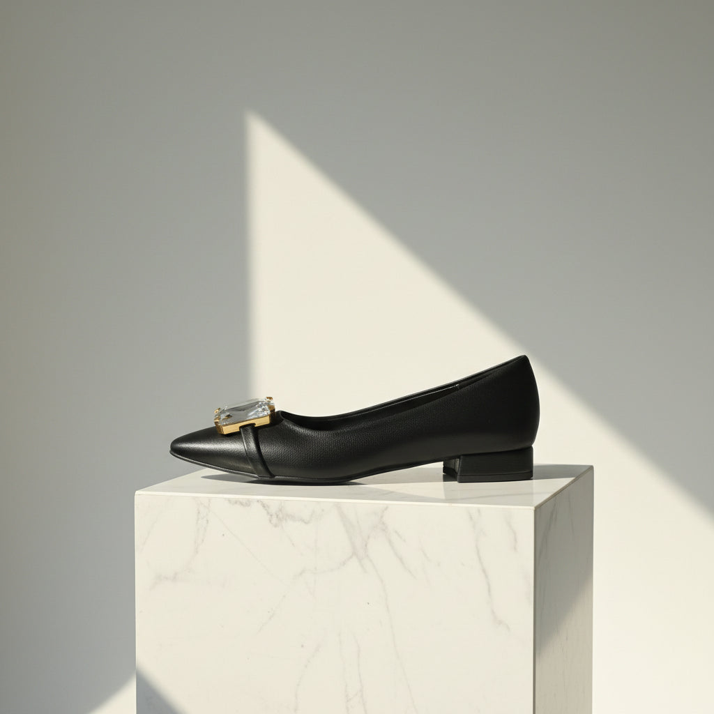 Black flat shoe with a decorative buckle on a marble pedestal against a gray background