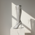 A tall silver peep-toe high-heel boot with a textured mesh-like surface and lace-up front, displayed on a white marble pedestal against a warm beige wall with dramatic diagonal sunlight and shadows.