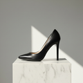 A single black leather high-heeled pump with a pointed toe, displayed on a white marble pedestal against a gray background with a sharp diagonal shadow.