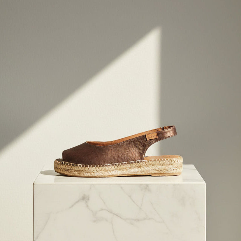 A single brown leather espadrille sandal with a braided jute platform sole, displayed on a white marble pedestal against a neutral gray background with a diagonal shadow.