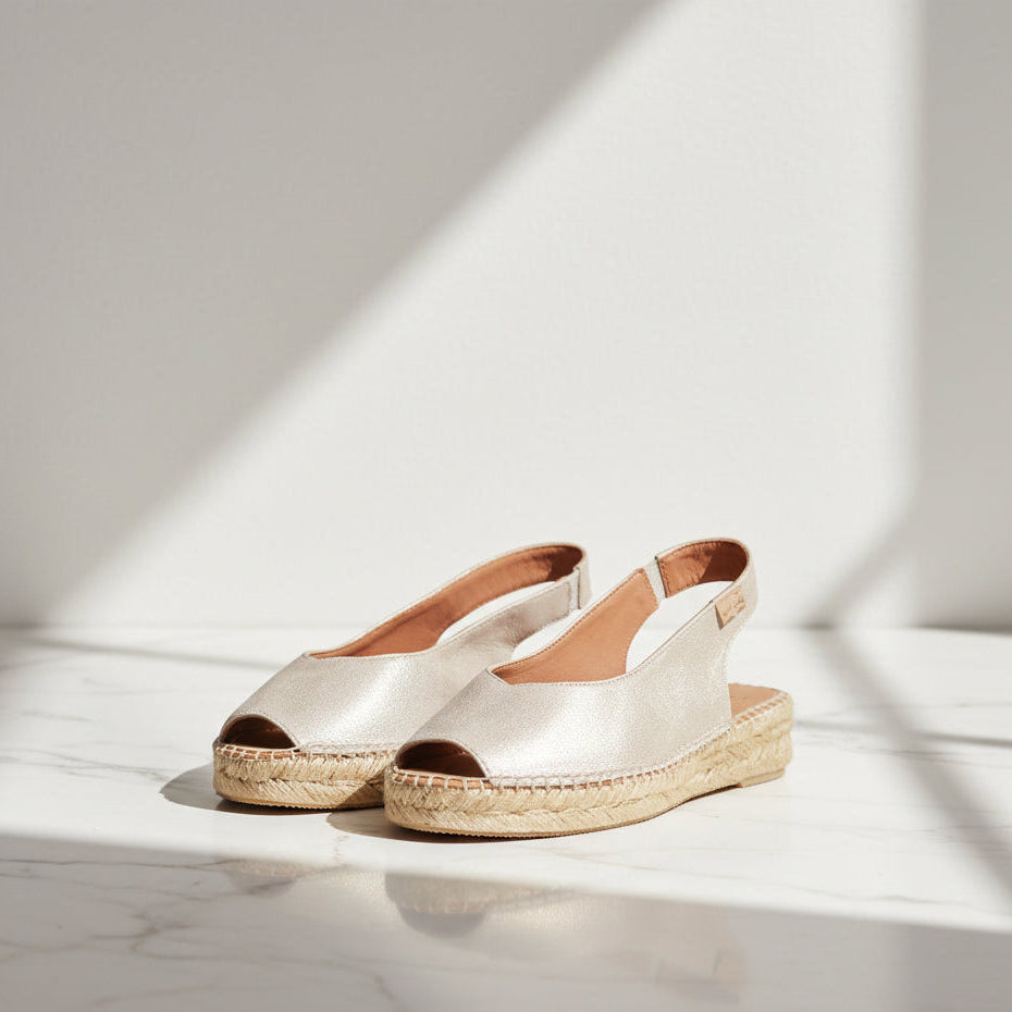 A pair of metallic champagne espadrille slingback shoes with woven jute soles, displayed on a white marble surface under soft diagonal sunlight.