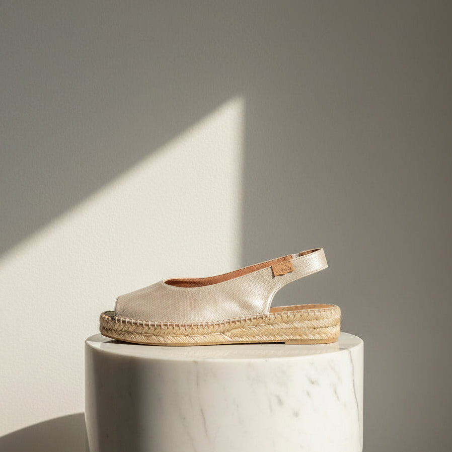 A single espadrille sandal with a metallic champagne-gold upper and a natural jute-wrapped platform sole, displayed on a white marble pedestal against a neutral gray wall with soft diagonal sunlight.