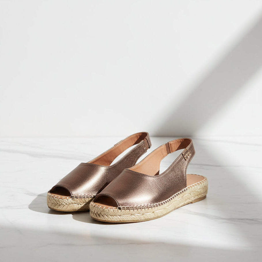 A pair of metallic rose gold espadrille wedge sandals with open toes and slingback straps, displayed on a light marble surface with soft shadows.
