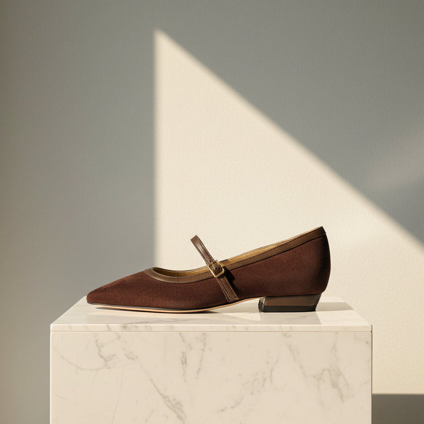 A single dark brown suede Mary Jane flat shoe with a pointed toe, a brown strap with a small gold-tone buckle, and a low block heel, shown against a light gray cardboard background.