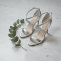 Silver high-heeled sandals on a marble surface with a sprig of eucalyptus.