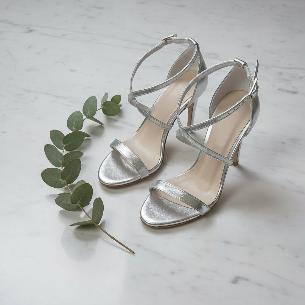 Silver high-heeled sandals on a marble surface with a sprig of eucalyptus.