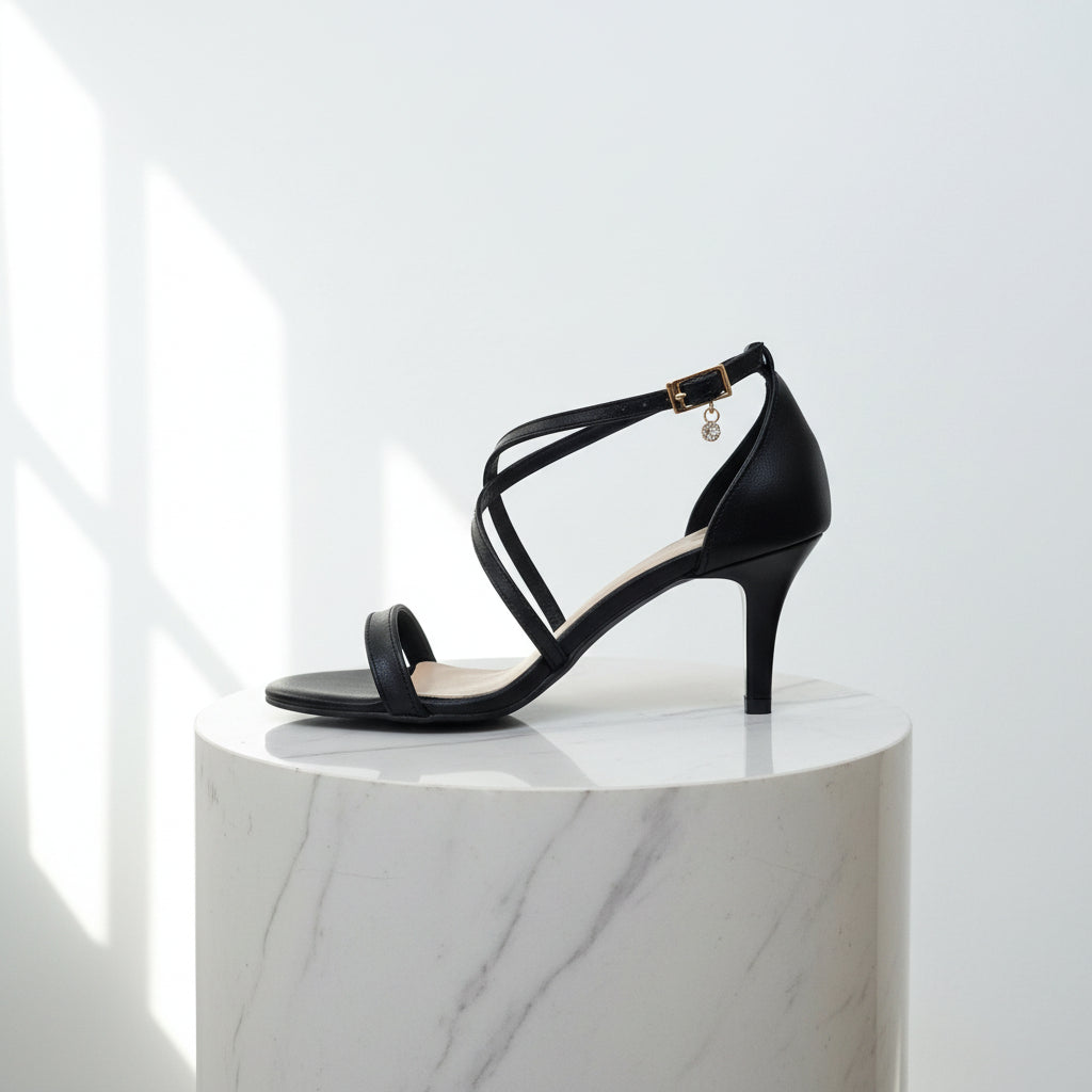Black high-heeled sandal with thin straps on a reflective surface.