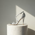 A single metallic silver high-heeled sandal with an ankle strap and open toe, displayed on a white marble cylinder pedestal against a warm gray wall with a diagonal shadow.