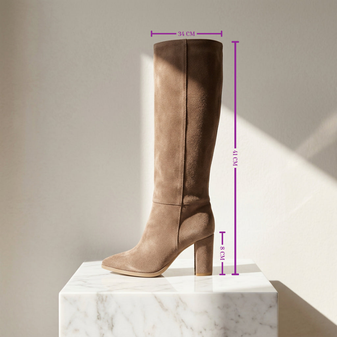A tall taupe-brown suede knee-high boot with a block heel and pointed toe, displayed on a white marble pedestal against a beige wall with purple dimension lines indicating height and width measurements.