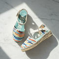 A pair of espadrille wedge sandals with colorful striped fabric uppers and jute-wrapped soles, displayed on a white marble surface with diagonal shadows.