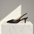 A single black leather slingback pump with a pointed toe and mid-height heel, displayed on a white marble pedestal against a gray background with a diagonal light shadow.