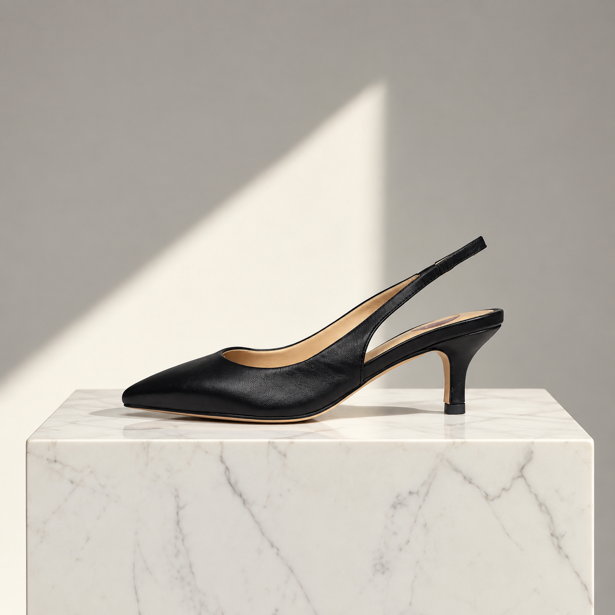 A single black leather slingback pump with a pointed toe and mid-height heel, displayed on a white marble pedestal against a gray background with a diagonal light shadow.
