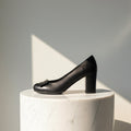 A single black leather high-heeled pump with a small bow detail on the toe, displayed on a white marble cylinder pedestal against a gray wall with a geometric shadow.