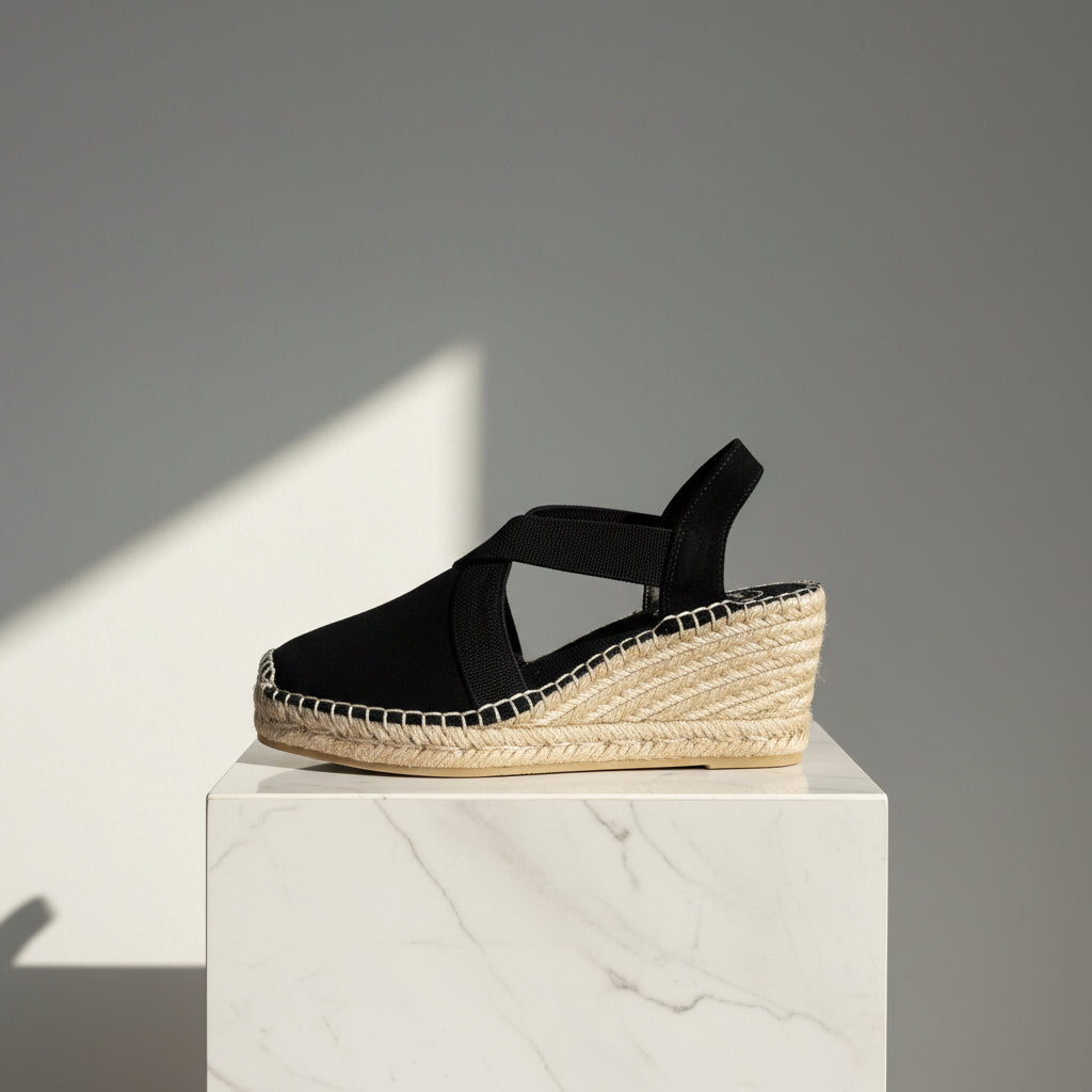 A single black wedge espadrille sandal with a braided jute platform heel and slingback strap, displayed on a white marble pedestal against a gray background with a diagonal shadow.