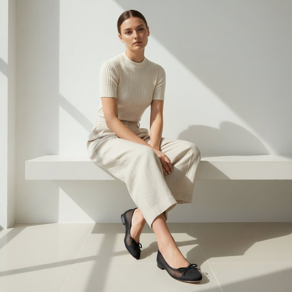 Woman sat on a minimalist wall bench wearing linen outfit and black mesh and bow flats. 
