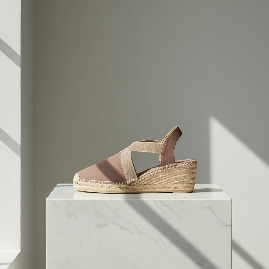 A single women's espadrille sandal in a muted dusty pink and beige colorway, resting on a white marble pedestal with dramatic window shadows.