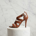 Brown high-heeled sandal with a silver buckle on a marble pedestal against a marble background