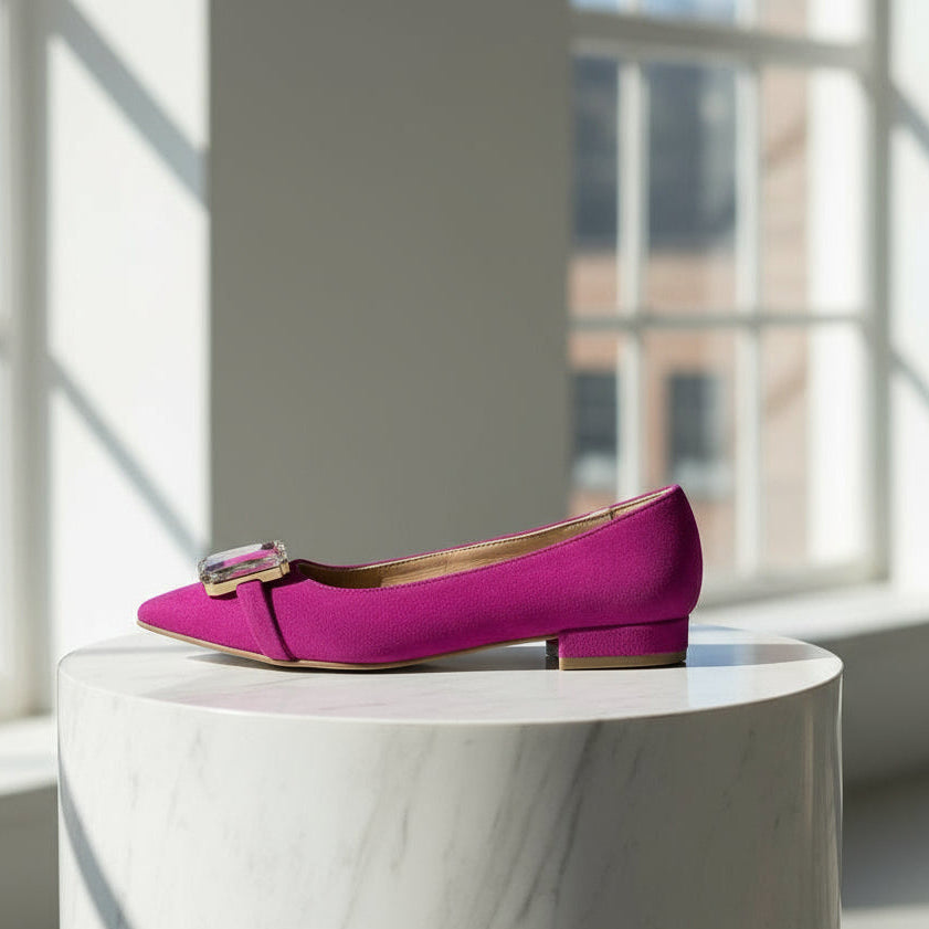 Hot pink suede ballerina with a gold buckle on a white marble surface in a bright room.