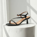 Black sandal with thin straps on a white pedestal against a light gray background
