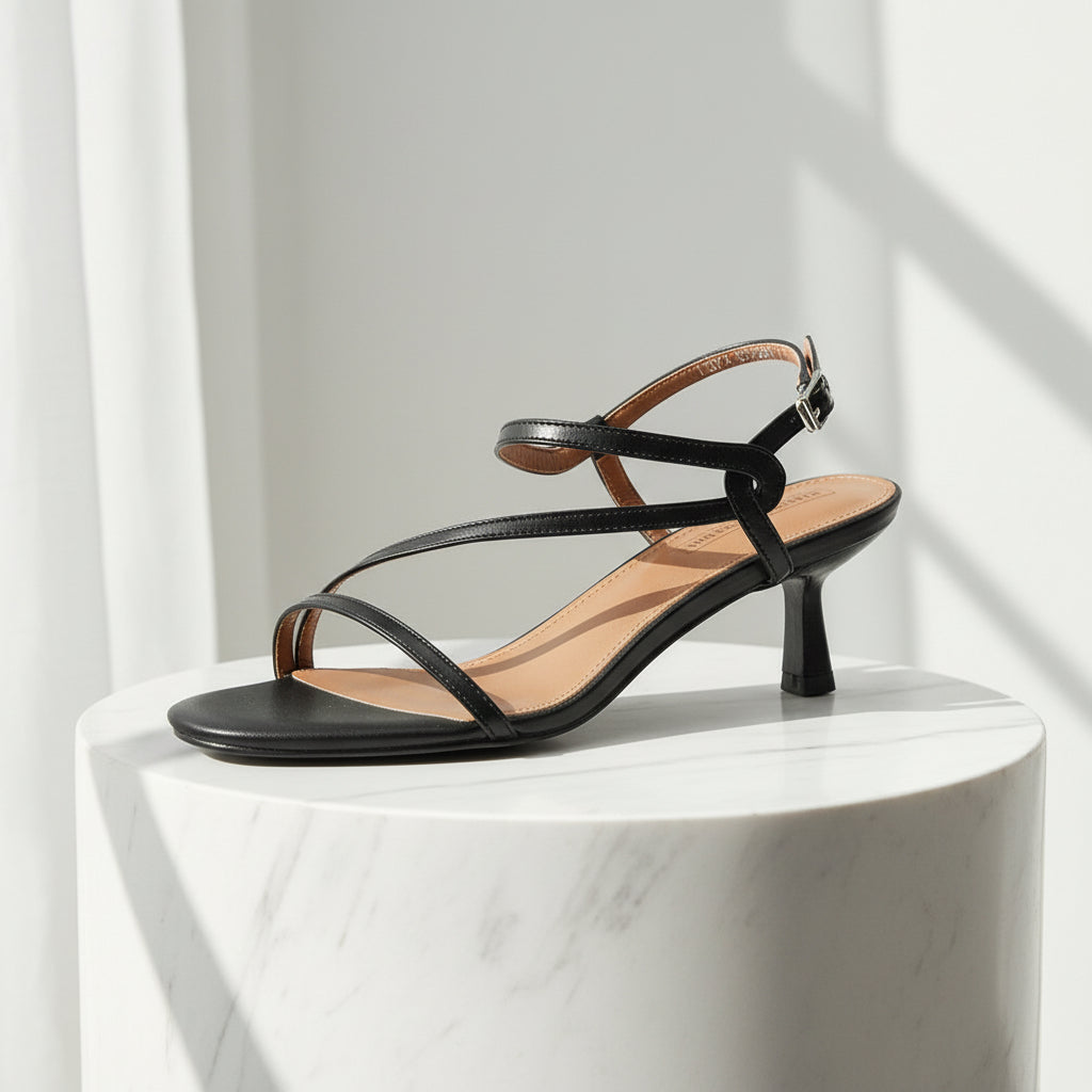 Black sandal with thin straps on a white pedestal against a light gray background