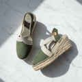 A pair of women's wedge espadrille sandals in olive green suede with beige woven straps and braided jute wedge heels, displayed on a white marble surface with diagonal shadows.