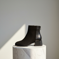 A black leather ankle boot with a low block heel and side zipper, displayed on a white marble cylinder pedestal against a neutral gray background with a diagonal shadow.